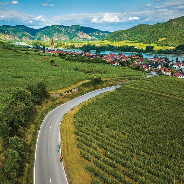 Aerial view of two people riding bicycles on a winding paved road through vineyards. There are houses, a river, and mountains in the background.