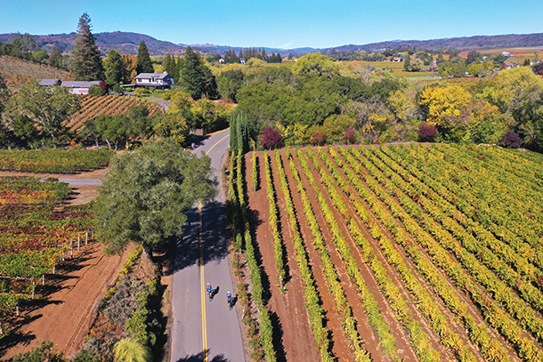 Aerial view of two cyclists on a winding road through California countryside and vineyards. There are houses and mountains in the background.