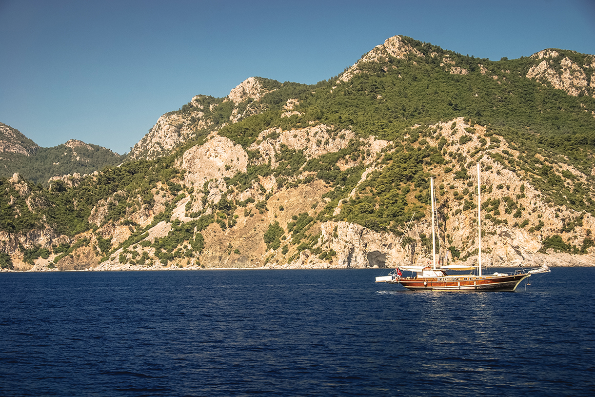 A traditional wooden yacht sailing in a large body of water with mountains in the background.