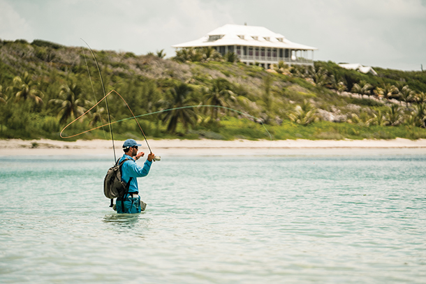 A man wearing blue fishing attire and a gray backpack, fly fishing off shore near The Delphi Club in the Bahamas. He is holding a fly rod and casting it out in the water.