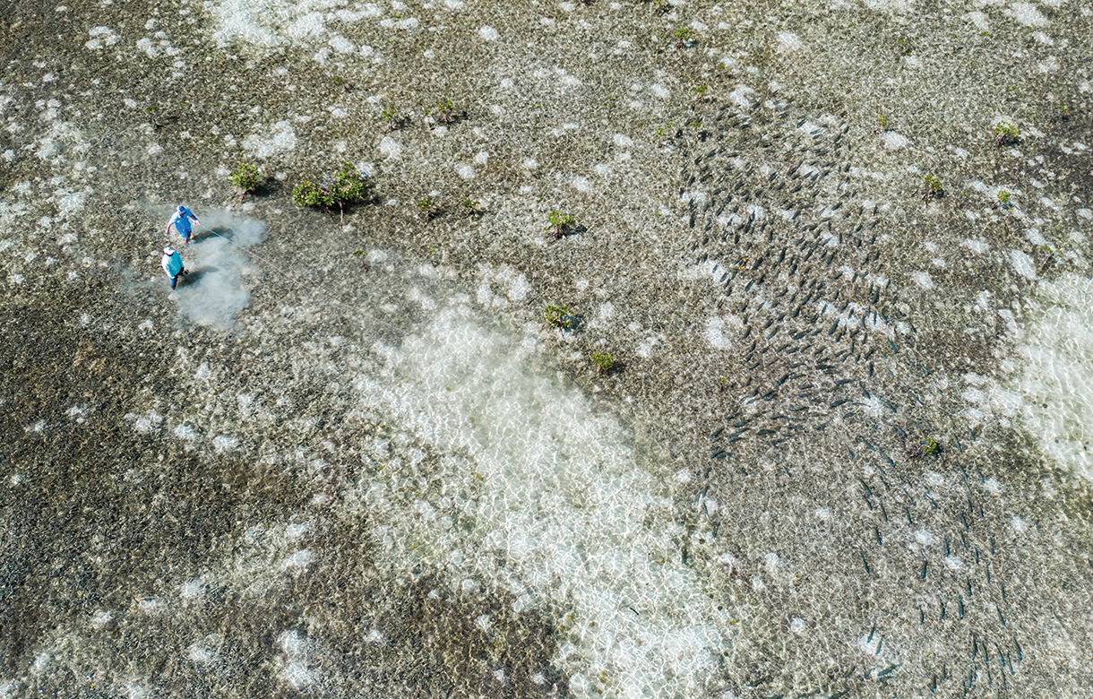 Aerial view of two people bonefishing in a vast area of shallow water in the Bahamian flats. There’s a long, light-colored sandbar that juts out into the water. There are also smaller, light-colored islands scattered throughout the water.