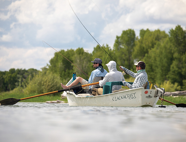 Three men fly fishing from a small white boat in a river. There are many trees surrounding the river. 