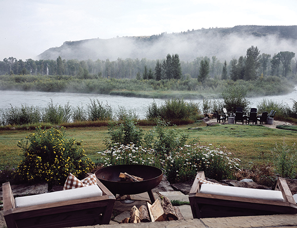 There are two wooden deck chairs next to a small fire pit at Idaho’s South Fork Lodge, with a view of a river in the background. There are many trees lining the bank. 