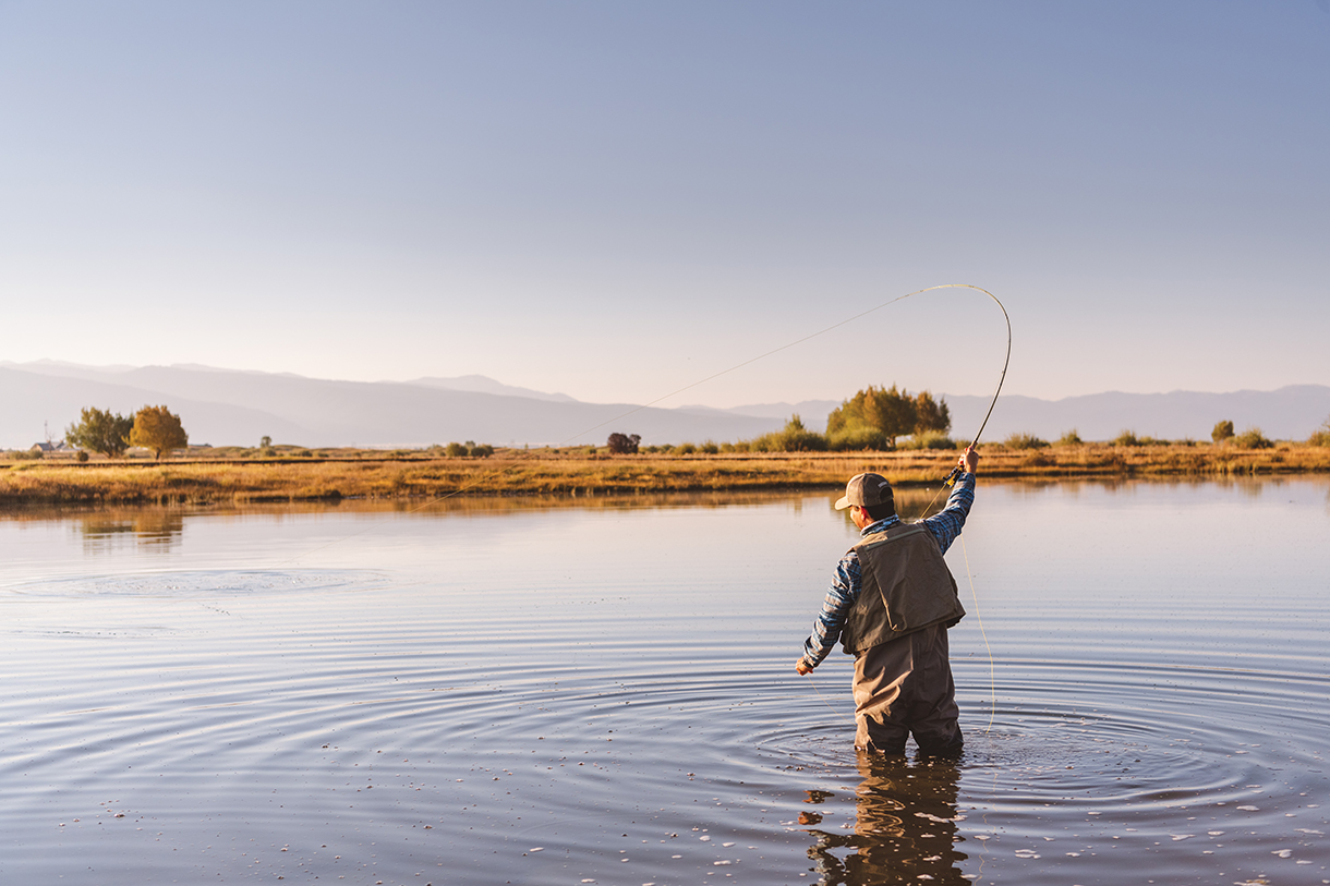 A man is fly fishing in a large, serene pond. He is standing in the middle of the pond, casting a fly rod. The water in the lake is calm and reflects the clear blue sky. There are mountains in the background.