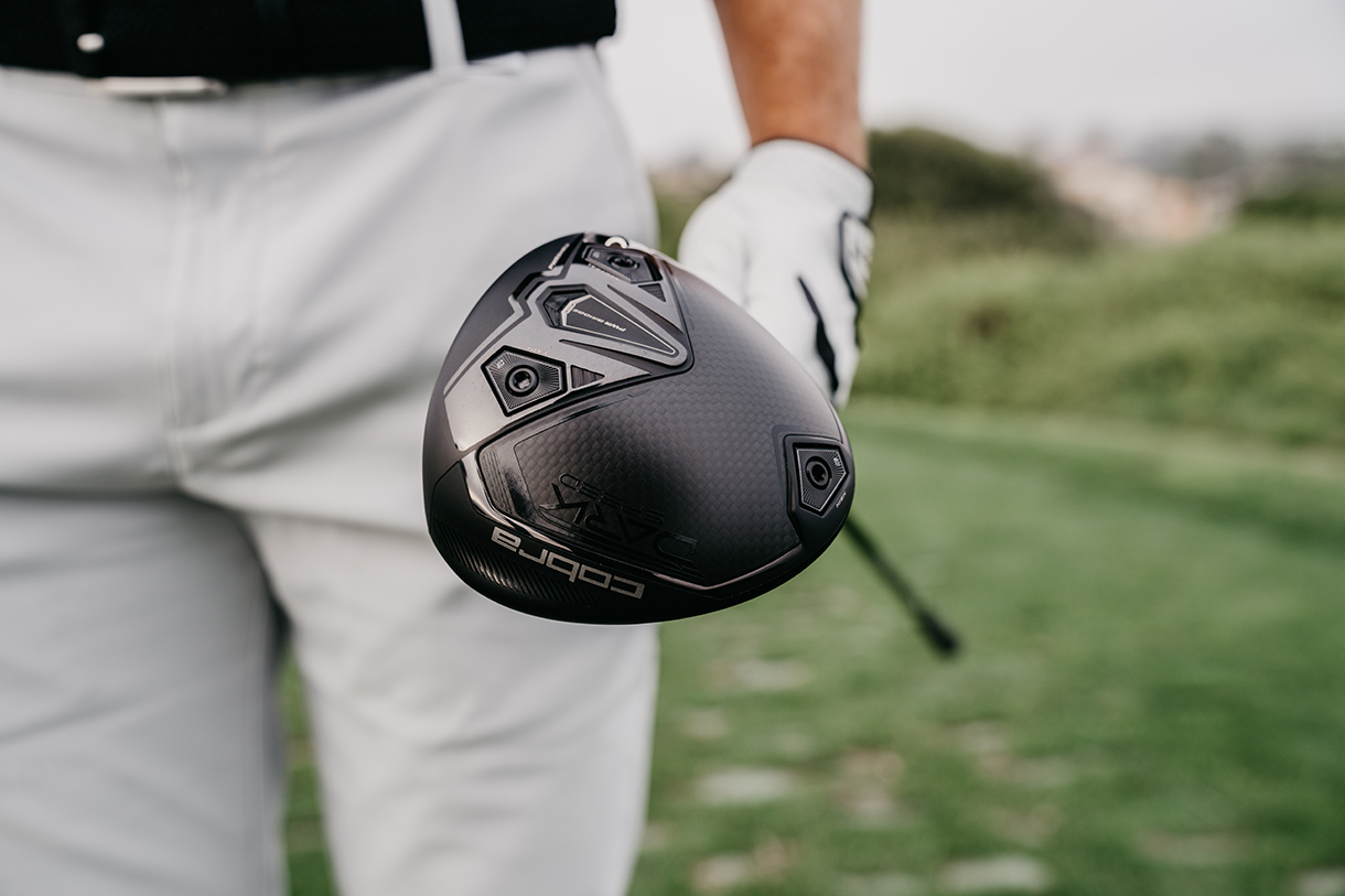 A golfer holding a Cobra DARKSPEED driver, showcasing the black clubhead with the Cobra logo and visible carbon fiber elements.