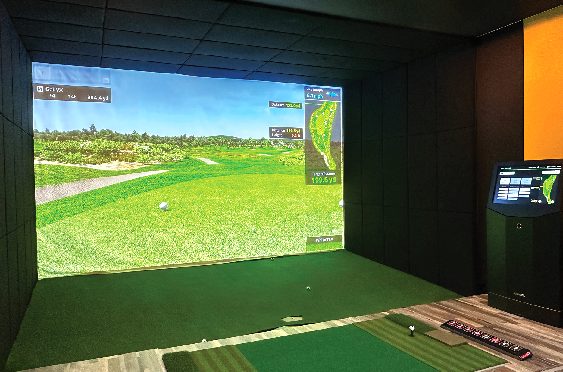 A golf simulator room with a large screen displaying a realistic golf course scene and a hitting mat in front. There is a computer monitor on the side showing golf data and statistics.