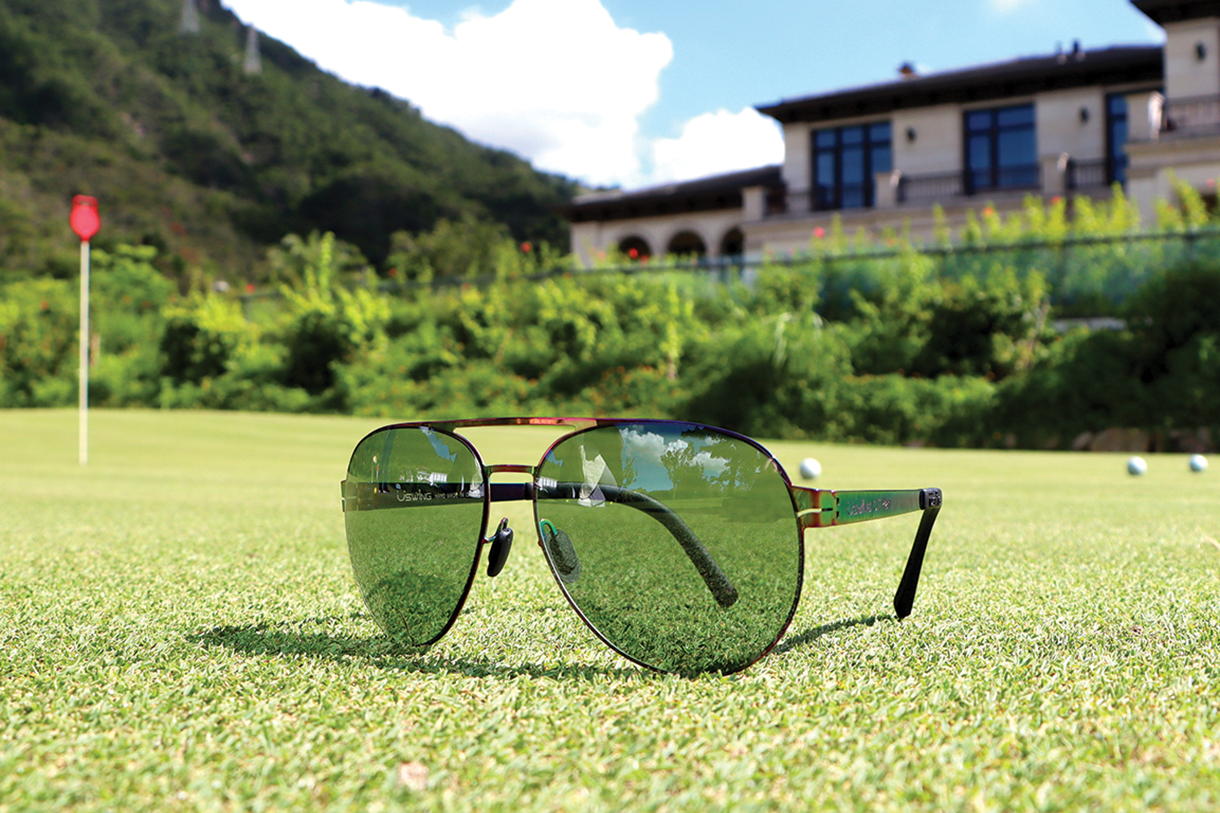 A close-up of a pair of black aviator-style Uswing Sunglasses resting on a green, with a golf course and a clubhouse in the background.