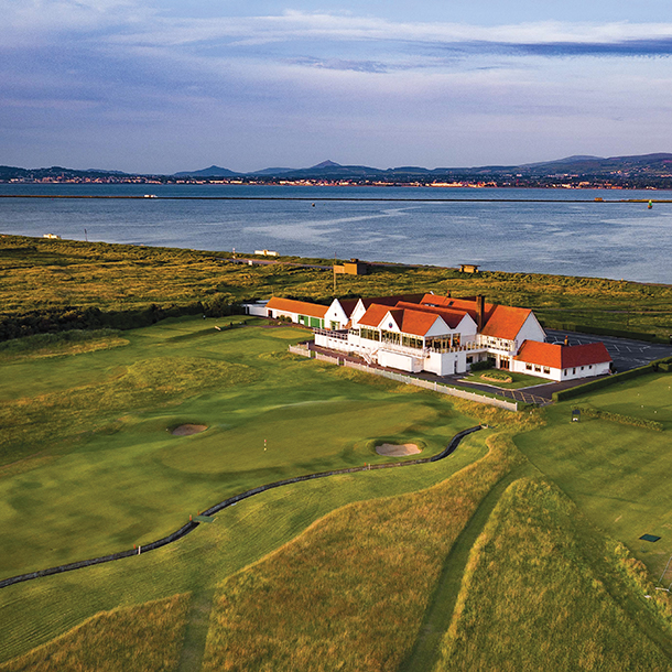 An aerial view of a white clubhouse surrounded by a golf course. There is a large body of water next to the golf course.