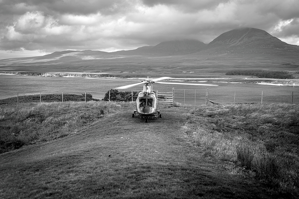 A black and white photo of a helicopter parked on a grassy hill with mountains in the background. The sky is cloudy and dramatic.