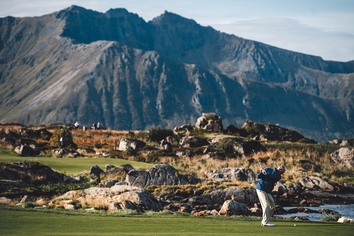 A golfer taking a swing on a scenic golf course with mountains in the background. The course is rocky and has a dramatic ocean view.