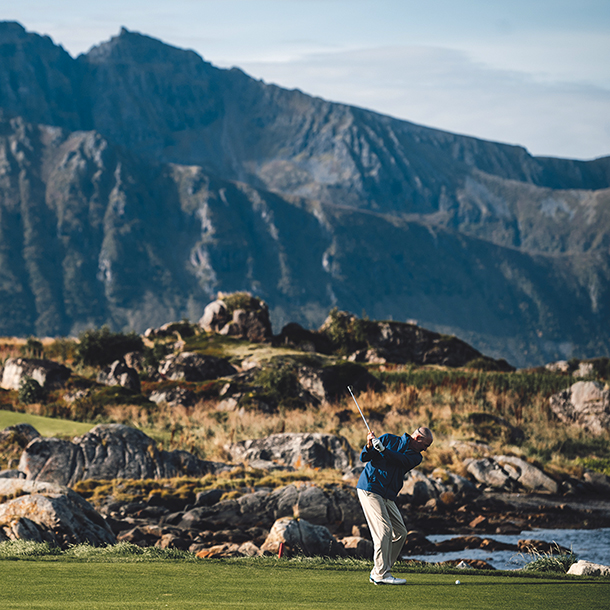 A golfer taking a swing on a scenic golf course with mountains in the background. The course is rocky and has a dramatic ocean view.