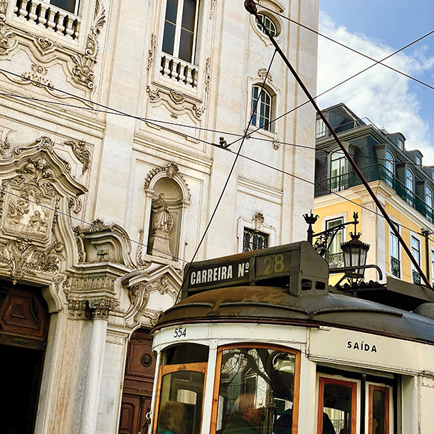 A vintage tram, labeled travels through a historic Lisbon street. The tram has a weathered exterior and is partially obscured by a building with ornate architecture and a statue. 