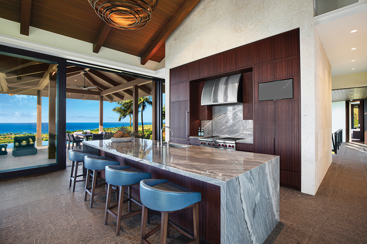 Interior of a modern luxury kitchen with a large island, marble countertops, and dark wood cabinetry. The kitchen has sliding glass doors leading to a covered patio with ocean views.