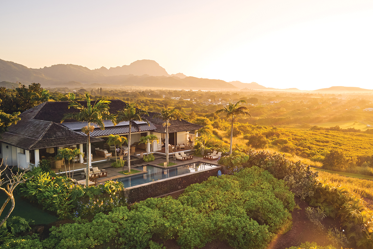 Aerial view of a modern luxury home with a pool, set against a backdrop of rolling hills and a distant mountain range at sunrise in Kauai.