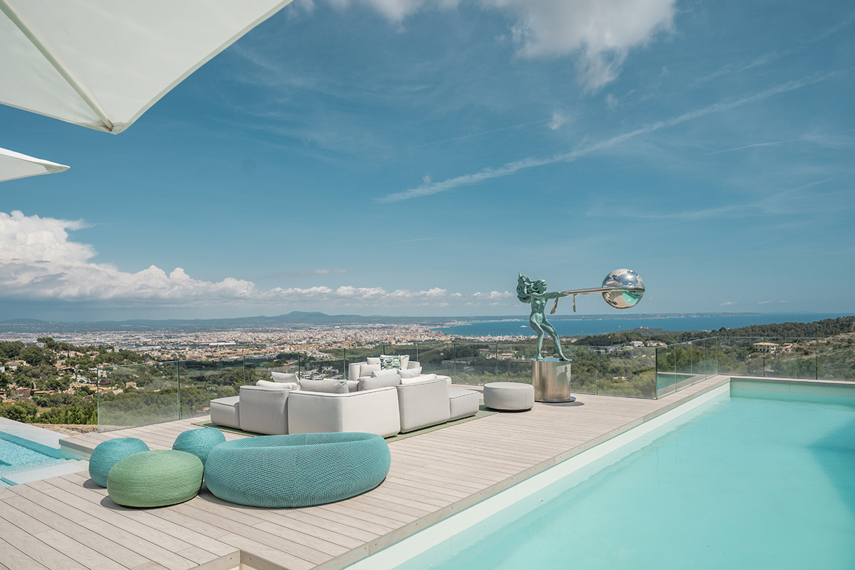 A luxurious outdoor terrace overlooking a panoramic coastal view. The terrace features a modern sectional sofa, armchairs, and ottomans in neutral colors, with a swimming pool in the foreground and a sculpture in the distance.