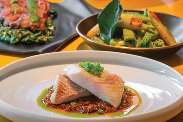 A plate of steamed sea bass with a red chili sauce in the foreground, served with vegetables in a bowl and grilled steak with greens on a separate plate.