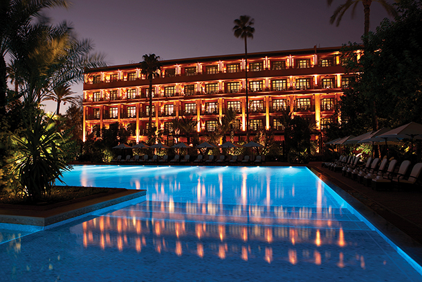 Exterior of La Mamounia Hotel in Marrakech, Morocco at night. The hotel is illuminated and reflects in the large, illuminated pool. Palm trees are visible on either side of the hotel.