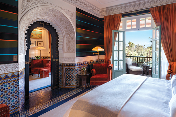 A luxurious hotel room with a king-size bed, ornate Moroccan tilework on the walls, and a view of a balcony overlooking a garden. The room has a sitting area with a sofa and a lamp, and the bed is made with white linens.