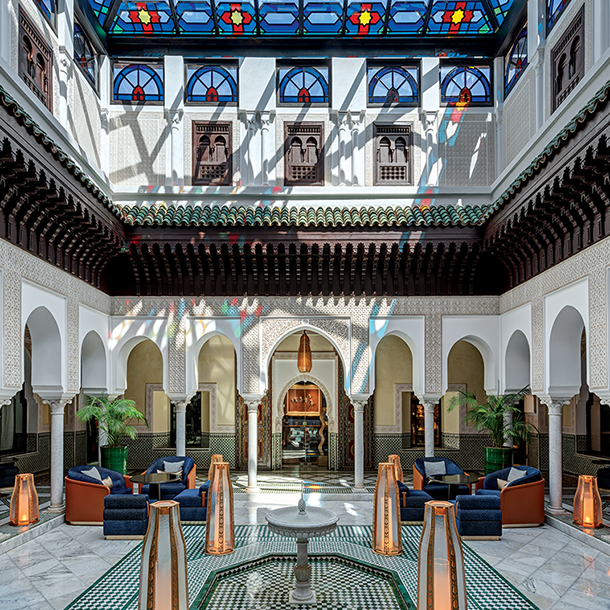 Interior shot of La Mamounia Hotel in Marrakech, Morocco. The image shows a large, open-air tea room and courtyard with a central fountain surrounded by arched columns and white walls. There are stained glass windows above and comfortable seating areas with blue armchairs and patterned rugs.