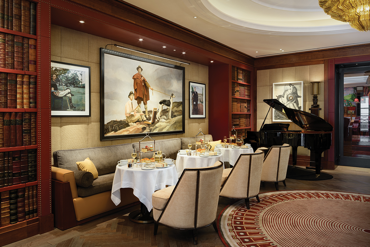 A cozy, elegant dining room with a vaulted ceiling. There are white tablecloths, plush armchairs, and a grand piano. Bookshelves line one wall, and a large painting hangs above a comfortable sofa.