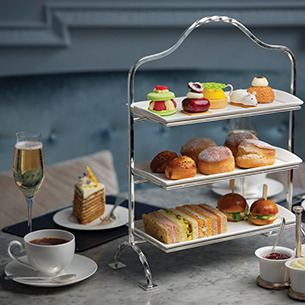 A three-tiered afternoon tea stand filled with various pastries, scones, and finger sandwiches. A cup of tea, a glass of champagne, and a slice of cake are also visible on the table.