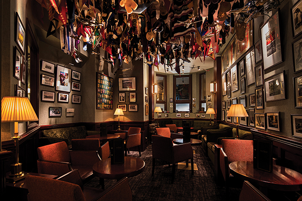 A cozy, dimly lit bar with a vintage atmosphere. The walls are adorned with photographs, memorabilia, and flags hanging from the ceiling. There are comfortable armchairs, small tables, and lamps throughout the room.