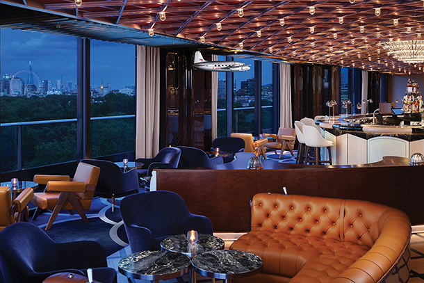 A stylish rooftop bar with floor-to-ceiling windows offering panoramic city views. The bar features a leather sofa, armchairs, and bar stools, as well as a hanging airplane model and a copper-toned ceiling.