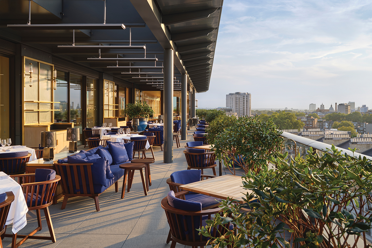 A rooftop terrace with outdoor seating, featuring blue armchairs, wooden tables, and potted plants. The terrace overlooks a cityscape with buildings and trees