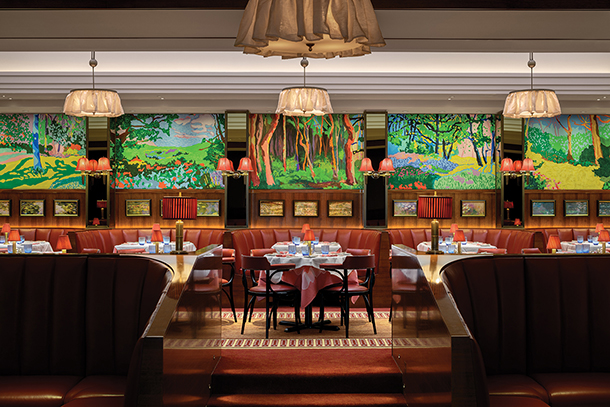 A stylish restaurant with red leather booths and round tables. The walls are adorned with colorful landscape paintings and there are hanging lamps with fabric shades.