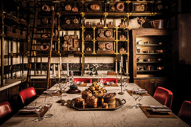 A cozy dining room with a long marble table set for a meal. There are red chairs, candles, and a variety of pies on a serving dish. The walls are lined with shelves filled with copper cookware and pie dishes.