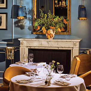 A cozy, elegant dining room with a fireplace, a large mirror, and a vase of flowers on the fireplace mantle. There are two round tables covered by white tablecloths with tableware on top in addition to upholstered armchairs.