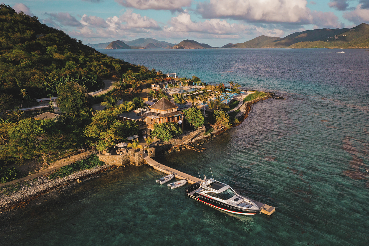 A white luxury yacht and two small boats docked at a wooden pier of a large property on a tropical island. Lush green palm trees, vegetation and ocean surround property with mountains visible in the background.