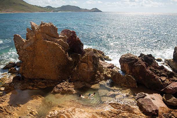 Two people swimming in a small rock pool along the shoreline. Waves are crashing against the rocks and the beautiful blue ocean is in the background with mountains in the far distance. 