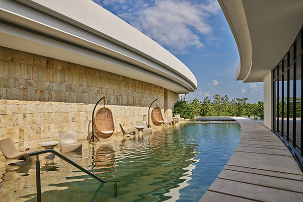 A long, narrow swimming pool that wraps around a building. There are several lounge chairs surrounding the pool. Beyond the deck, there is a wall of windows with a view of lush green palm trees and the surrounding area.