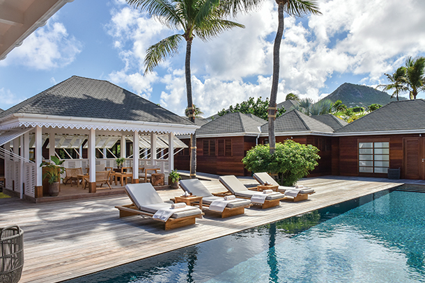 A large outdoor swimming pool surrounded by a wooden deck at a tropical resort. There are several lounge chairs on the deck and a gazebo in the background.