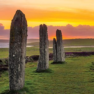 A row of four Neolithic stones which are large and upright located in a large field of grass at sunset. The sky is a gradient of orange, pink, and purple. There is a body of water and mountains in the distance.