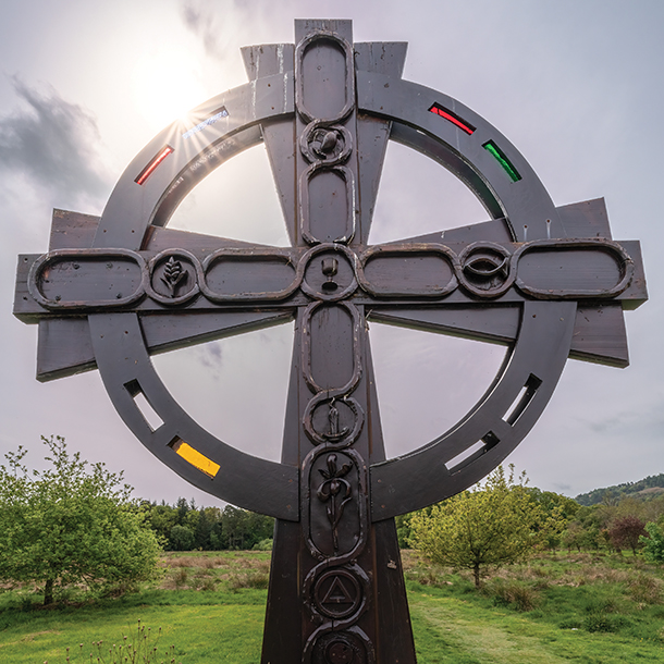  Close up of a large, intricately crafted Celtic cross. The upper part of the cross has a circular shape, and the details of the carvings are focused on Christian symbols. It is in a field of rolling hills and green grass.