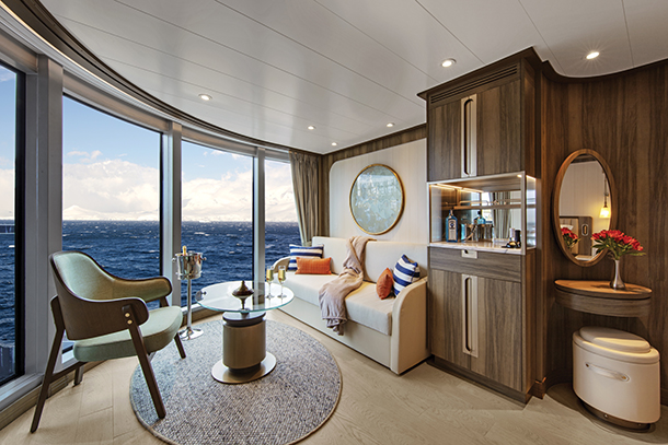 A stylish and contemporary seating area inside a luxurious suite aboard the Seabourn Venture. There is a sofa, armchair, coffee table, bar and large floor-to-ceiling windows that overlook the ocean.