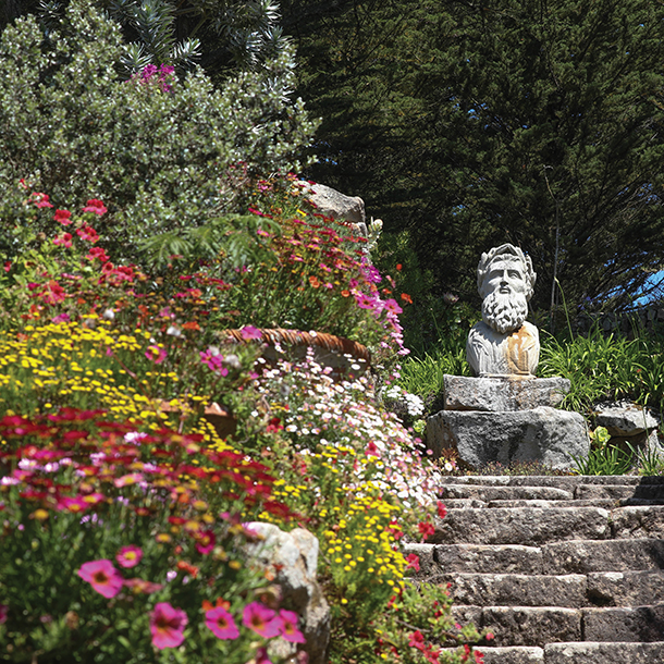 A beautiful garden with a. variety of colorful flowers planted. There is a statue of a man’s head placed on top of a large rock and an aged stone staircase that leads up to it.