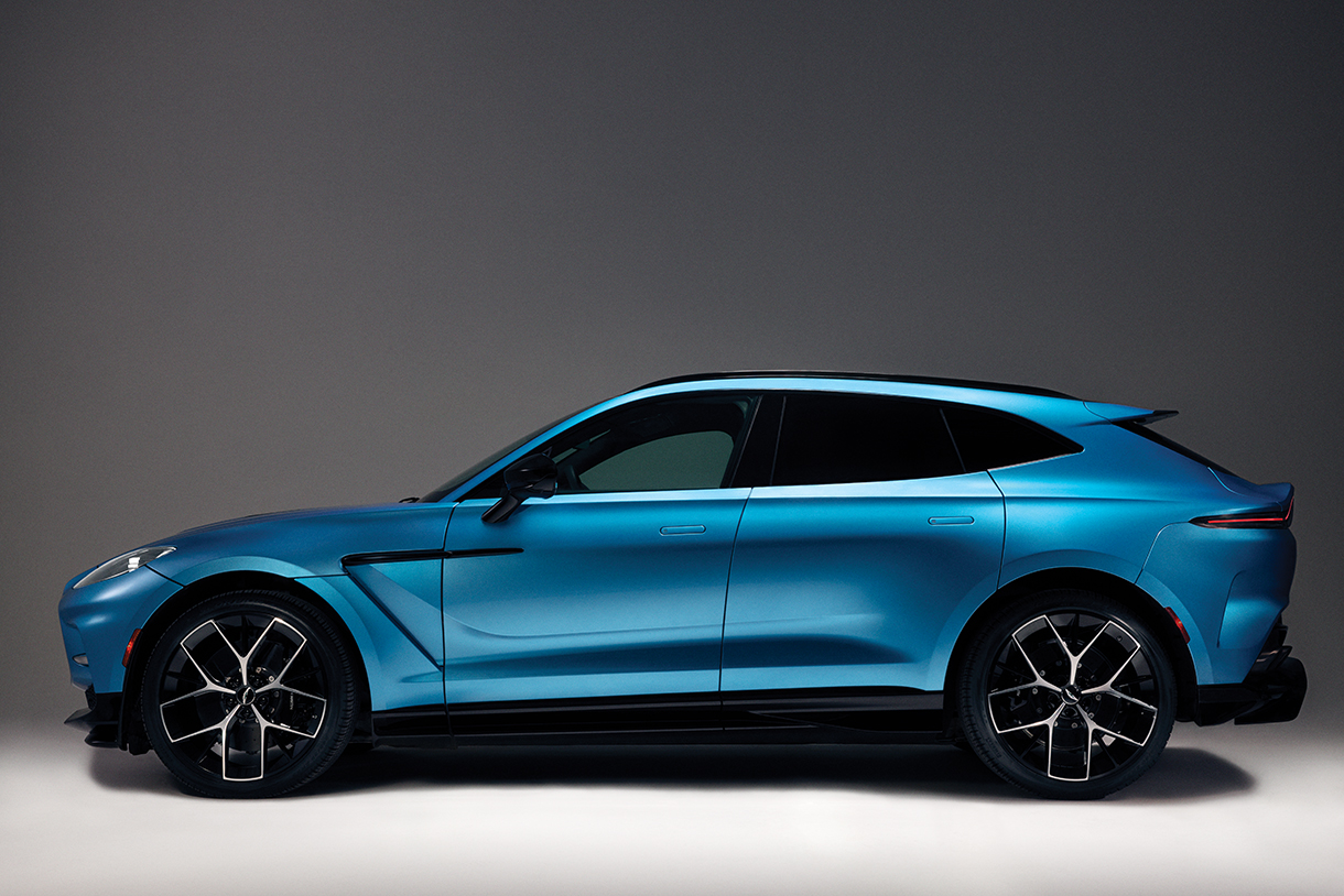 A side view of a blue Aston Martin DBX SUV parked in a studio or showroom. The SUV has black wheels and a luxurious, modern, and sleek design.