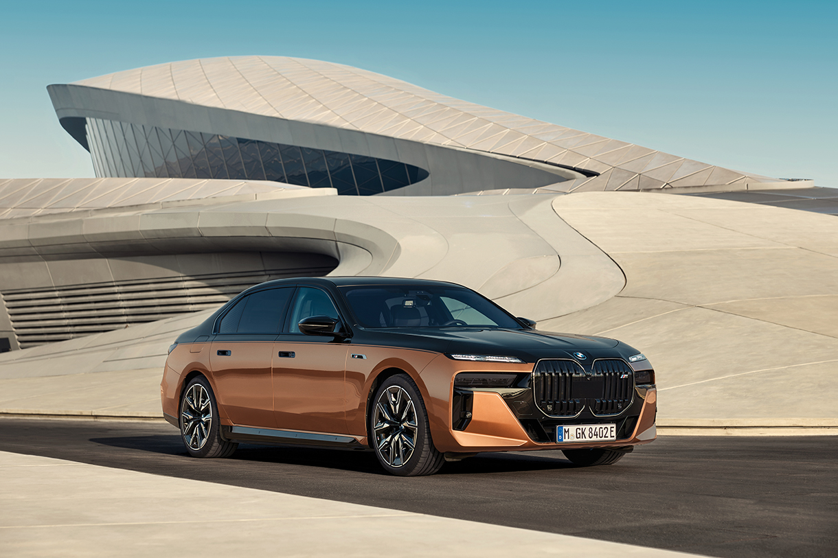 An orange and black BMW i7 M70 Electric SUV parked in front of a futuristic looking building or parking structure