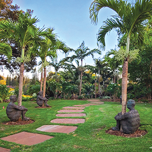 A serene garden path lined with stepping stones, leading to a cluster of sculptures nestled among palm trees and lush greenery.