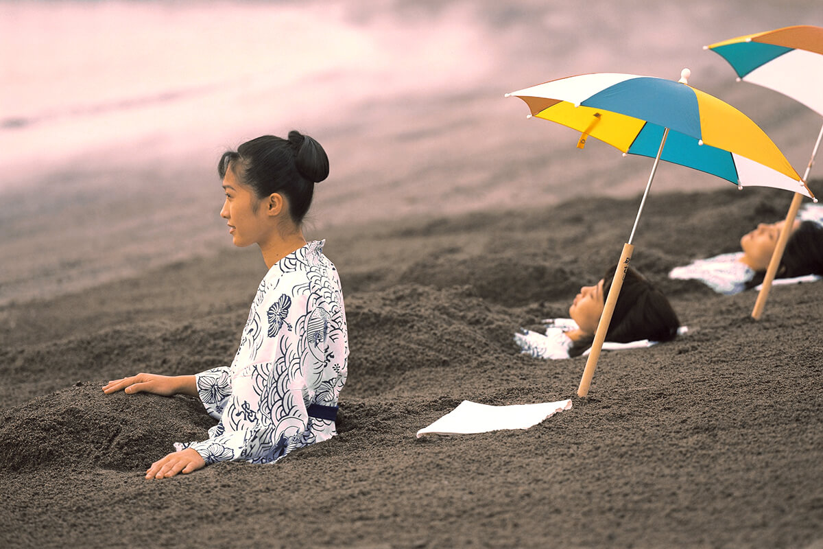 People lying down on their back on a sandy surface, covered from the neck down in what appears to be black sand. The person's head and hands are visible and they are reasting their heads on a white towel. 