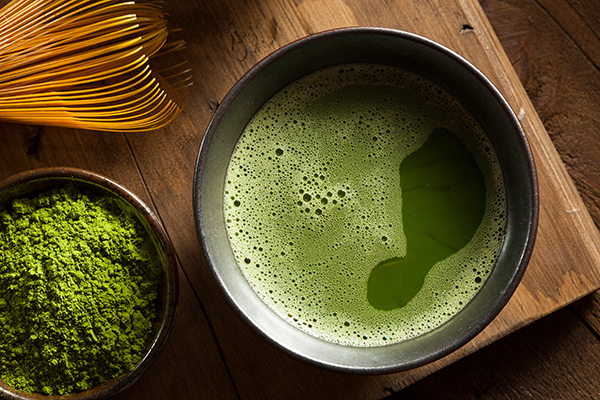 A cup of Japanese green tea and loose tea leaves. The background is a light wood table and there is a white ceramic teapot in the background with a wooden lid. 