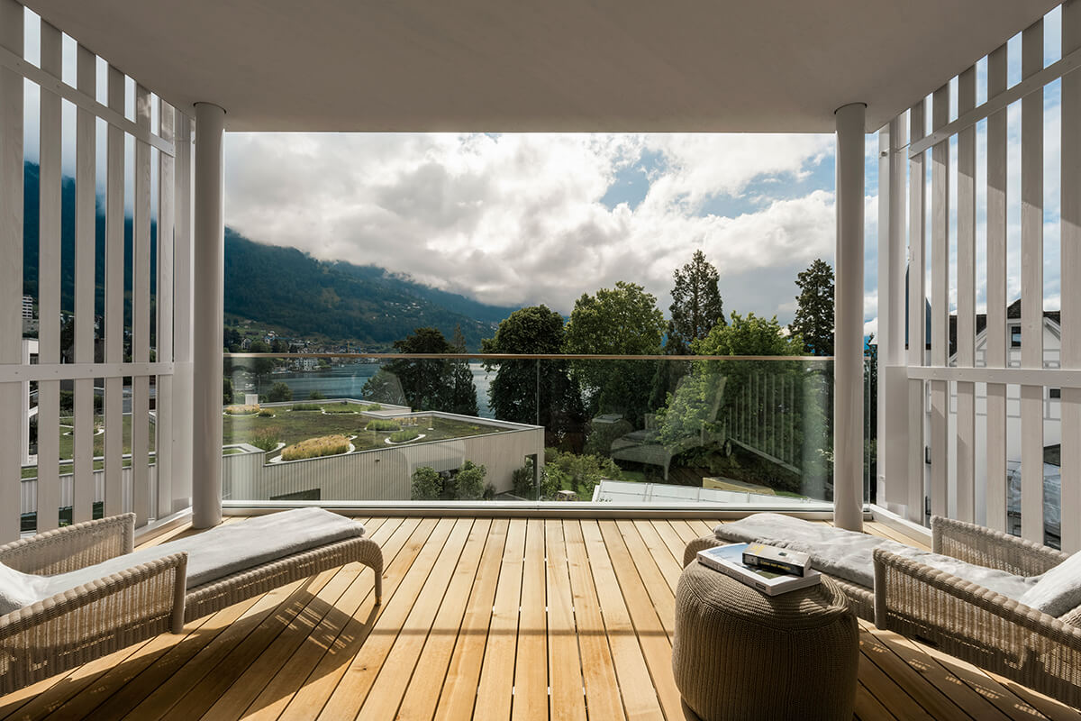 A beautiful and serene view from the wellness retreat at Chenot Palace Weggis of a mountainous landscape with a blue lake. The mountains are covered in green trees, and there is a small village or town in the distance. The image is taken from a high point from a terrace with some outdoor furniture.