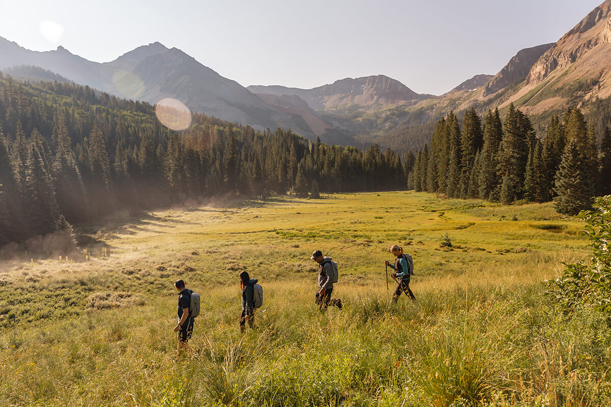 A group of hikers walking on a dirt path with a mountain range in the background. The hikers are all dressed in athletic wear and carrying backpacks, with some using trekking poles.