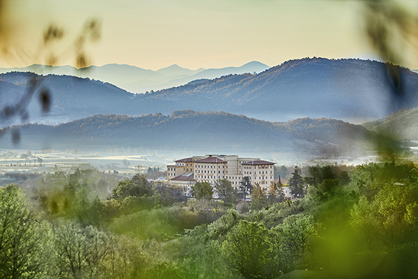 An exterior view of Palazzo Fiuggi, a wellness retreat located in the Italian countryside. The building is a historic stone palace with beige and brown hues, and it is surrounded by green trees and bushes. 