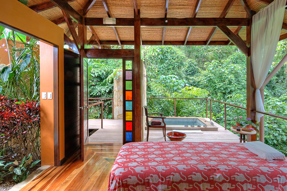 An outdoor spa treatment pavilion surrounded by lush greenery in a rainforest setting. The pavilion has a thatched roof and is supported by wooden pillars. In the foreground, there is a small pool of water. Inside the pavilion, there is a massage table adorned with a patterned linen. The image conveys a sense of tranquility and relaxation in a natural, tropical environment.