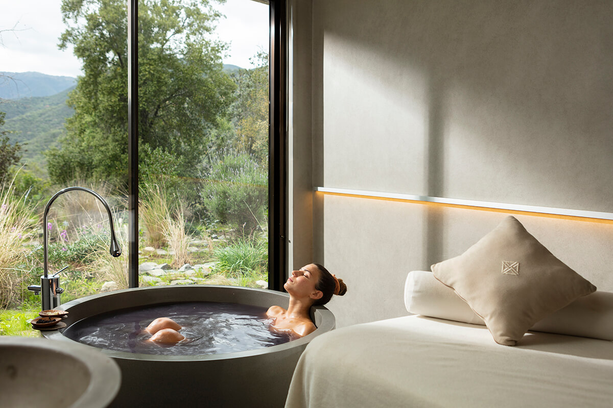 A luxurious spa bathroom with dim lighting and warm tones. The centerpiece of the room is a large circular soaking tub filled with steaming water.  In the background, there is a floor-to-ceiling glass window offering a breathtaking view of mountains and forest outside.