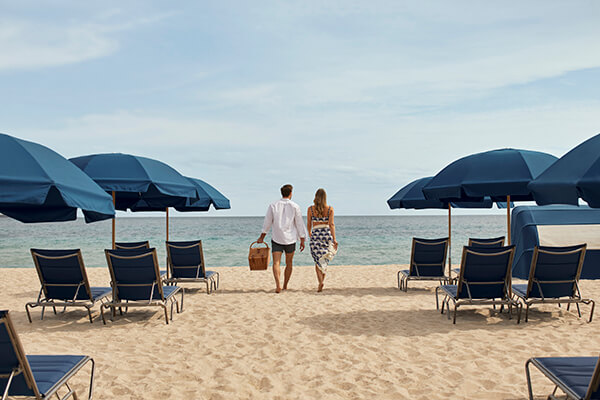 A beautiful beach setting with clear blue water, white sand and blue skies. A couple holding is hands walking towards the shoreline surrounded by multiple beach chairs and blue umbrellas. 